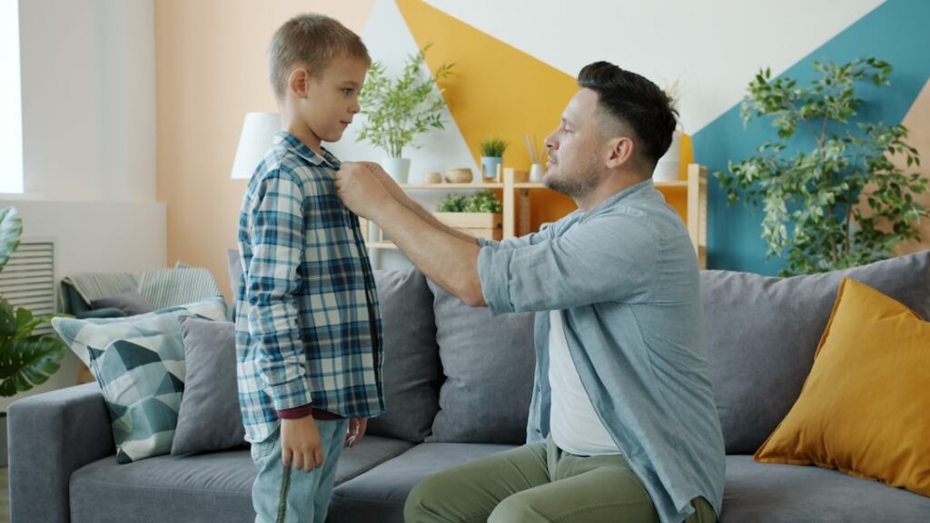 Caring father is helping little boy with clothing buttoning shirt talking indoors at home showing love and care. Family relationship, people and lifestyle concept.