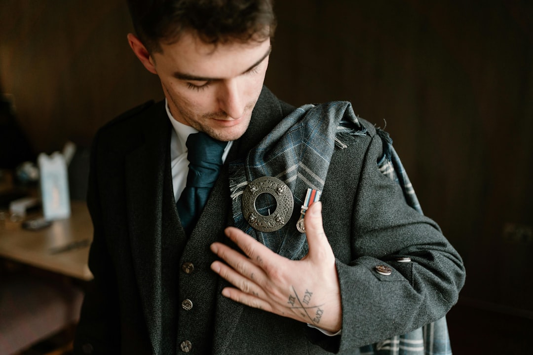 The groom stands proudly, adorned in a traditional Scottish kilt ensemble that reflects his heritage. The kilt, crafted from his family's tartan, showcases a rich pattern of deep greens and navy blues, symbolizing his clan's history. Paired with the kilt is a crisp white dress shirt and a charcoal grey waistcoat, adding a touch of modern elegance to the classic attire. A black leather sporran with silver detailing hangs at the front, serving both functional and decorative purposes.