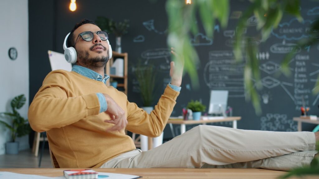 Middle Eastern guy in casual clothing is enjoying music in wireless headphones singing moving arms dancing at work. Youth culture and people concept.