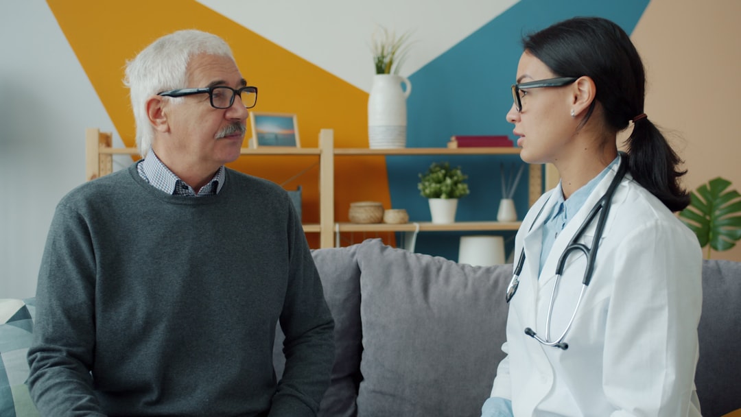 Young female doctor in white gown and glasses is talking to senior man patient at home expressing care and support. Medicine and old people concept.
