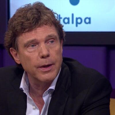 John de Mol als gast aan tafel in een televisieprogramma, met het Talpa-logo achter hem