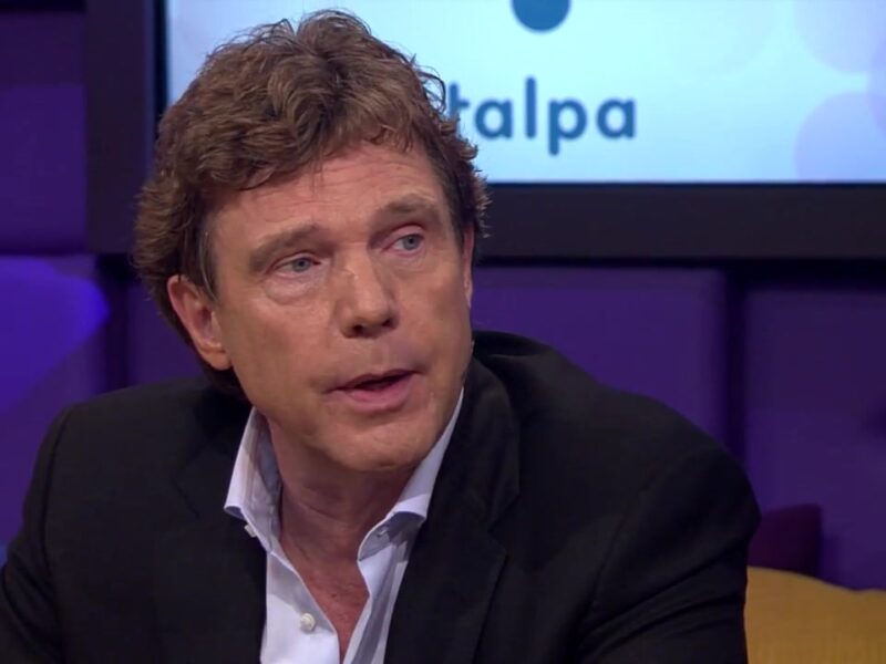 John de Mol als gast aan tafel in een televisieprogramma, met het Talpa-logo achter hem
