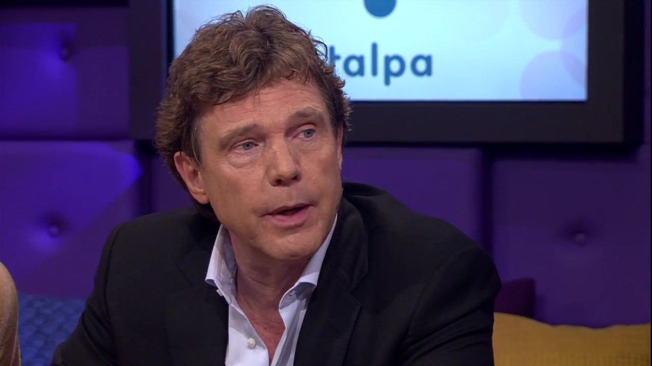 John de Mol als gast aan tafel in een televisieprogramma, met het Talpa-logo achter hem